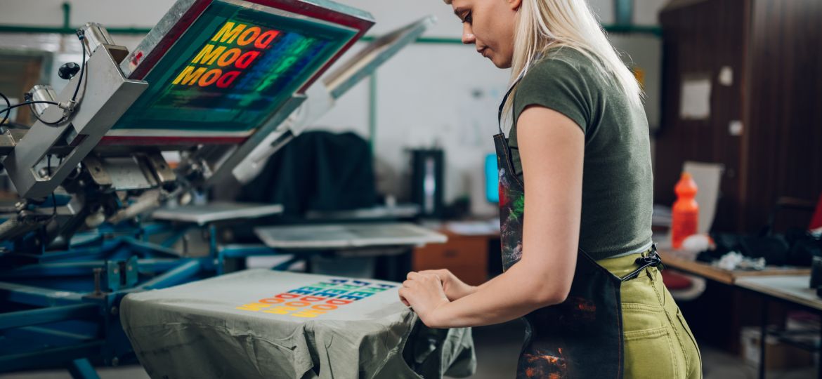 Print shop worker taking screen printed t-shirt of machine at facility