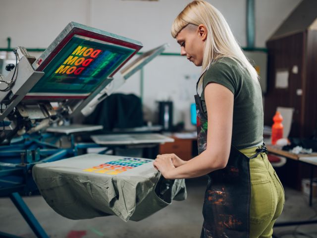 Print shop worker taking screen printed t-shirt of machine at facility