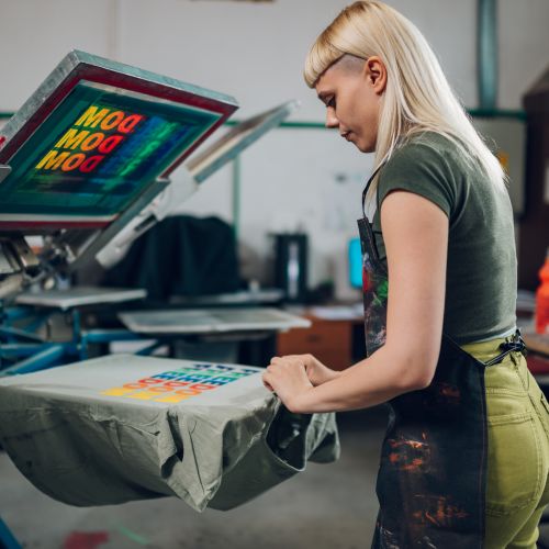 Print shop worker taking screen printed t-shirt of machine at facility